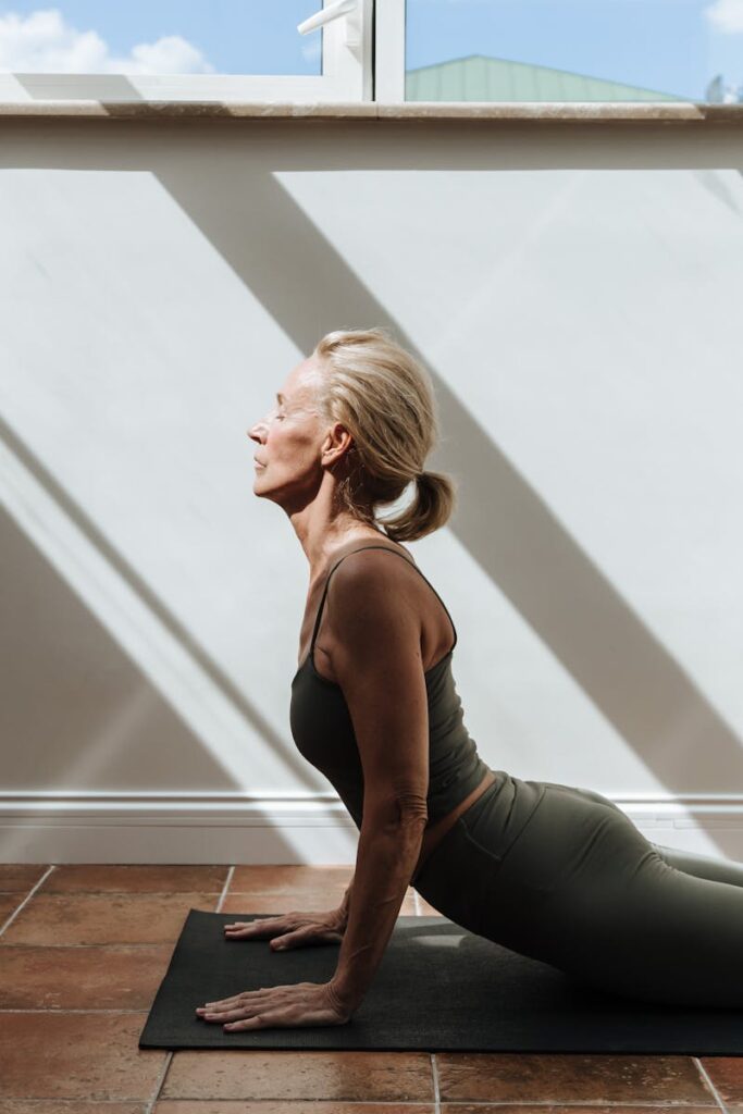 Elderly woman practicing yoga indoors with sunlight streaming through windows, embodying health and fitness.