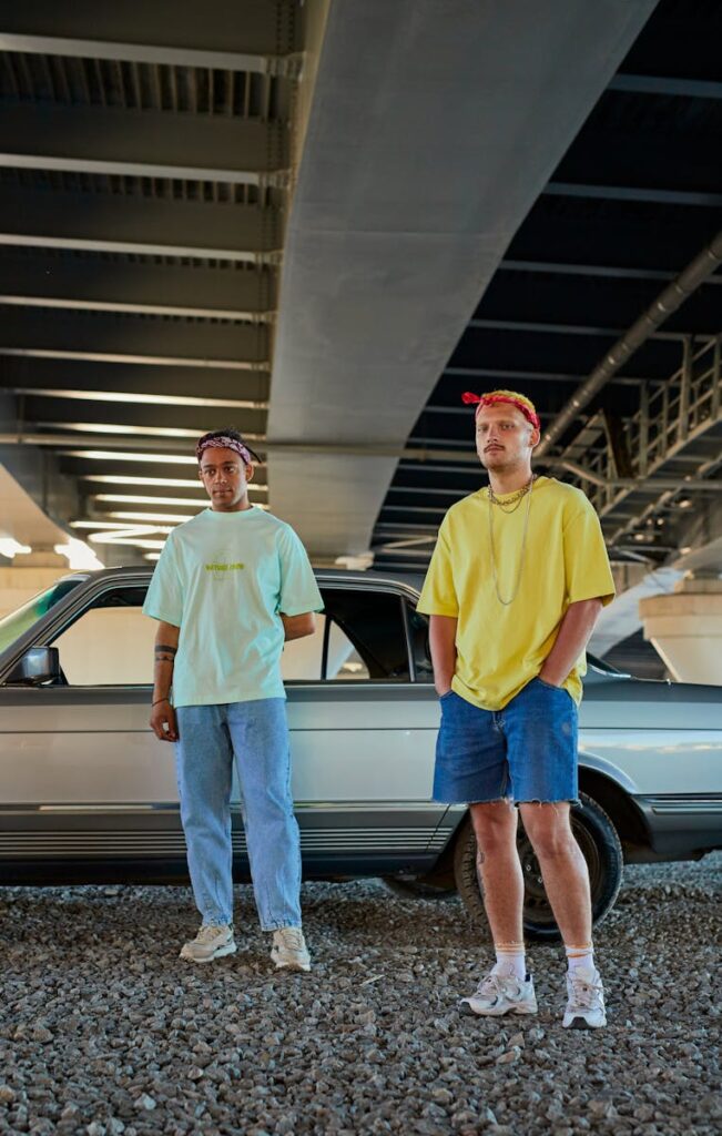 Two men in streetwear under a city bridge with a car, embodying urban fashion.