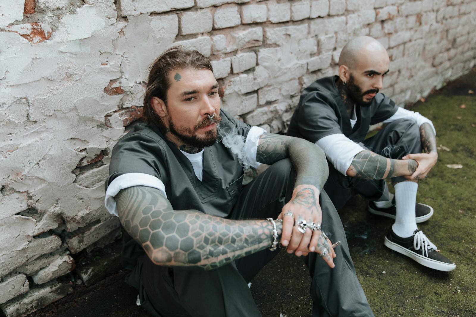 Two tattooed men in prison uniforms sit against a brick wall, exuding a gritty and rebellious vibe.