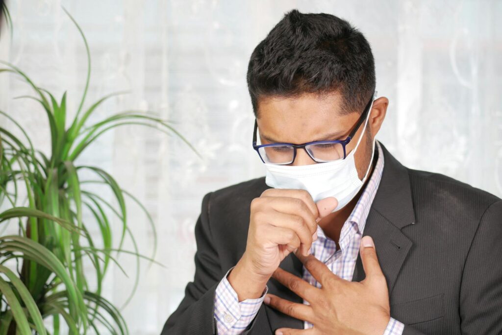 Asian man coughing in a black suit with eyeglasses and face mask indoors.