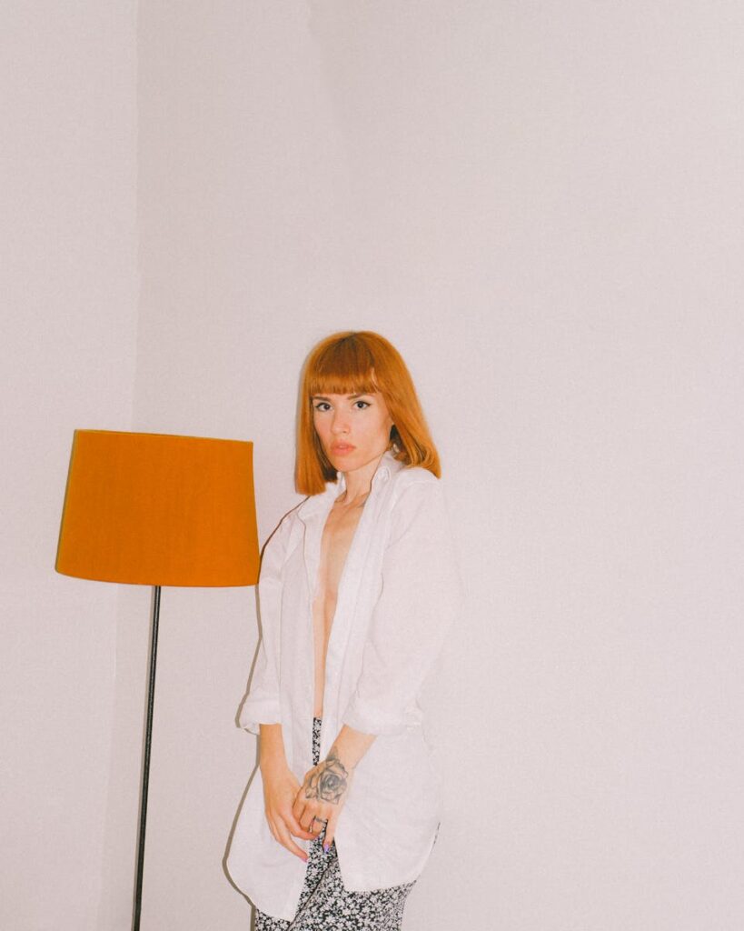 Caucasian woman with red hair posing indoors next to a lamp.