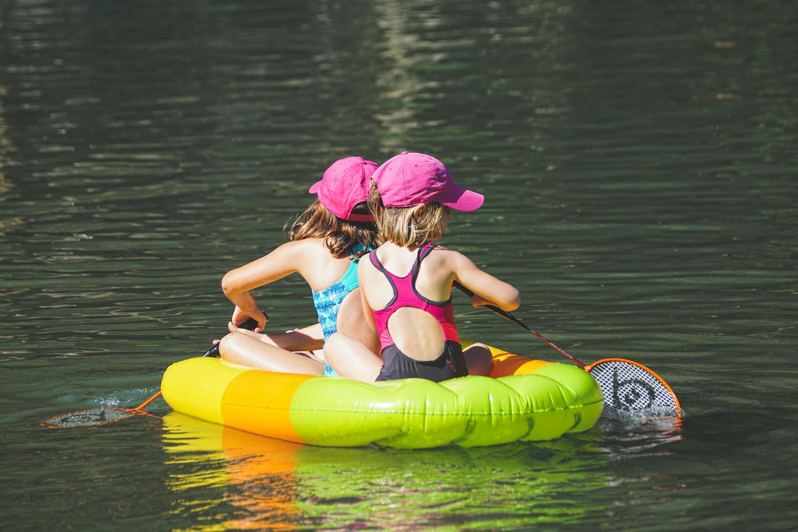 Two young girls paddle a colorful inflatable raft on a sunny lake day.