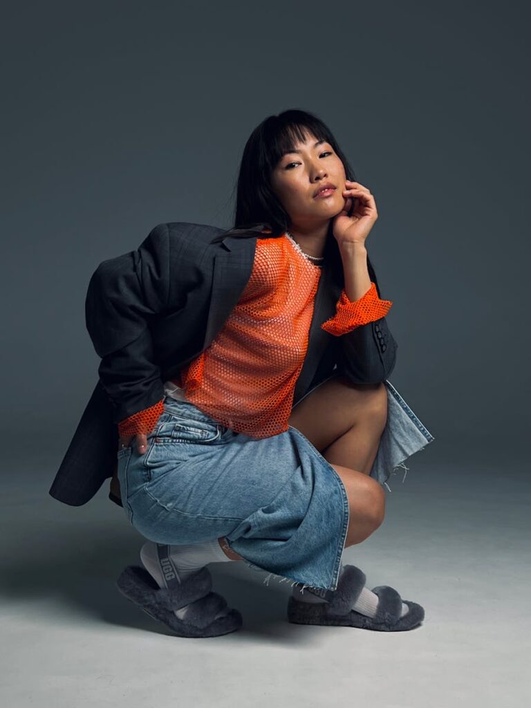 Fashionable Asian woman posing in studio wearing orange mesh top and denim skirt.
