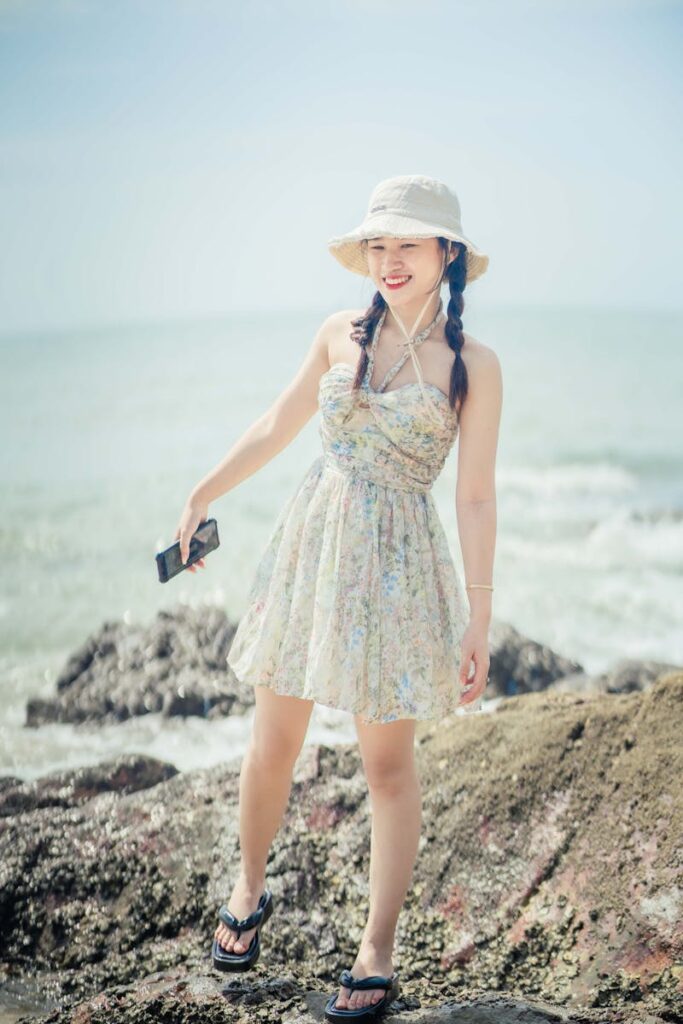 A young woman in a floral dress and hat smiles by the beach during a sunny day.