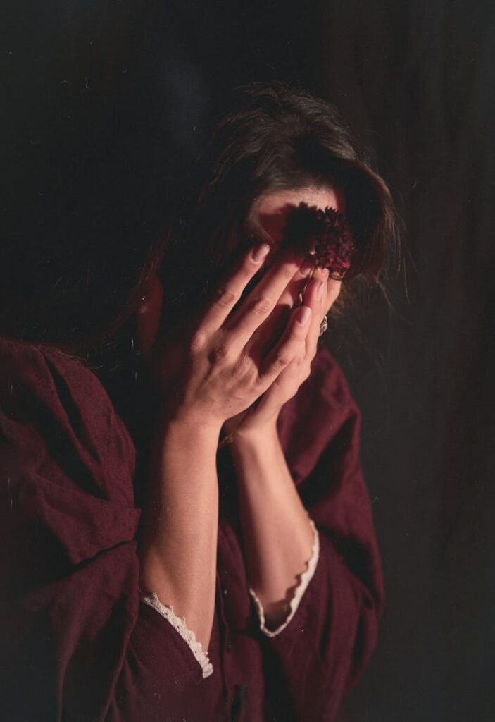 A woman in dark clothing holds her head in a mysterious, moody setting.