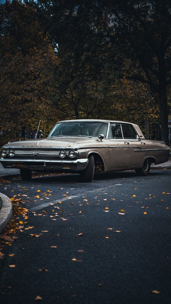 Classic Mercury car on a winding autumn road with fallen leaves and a forest backdrop.