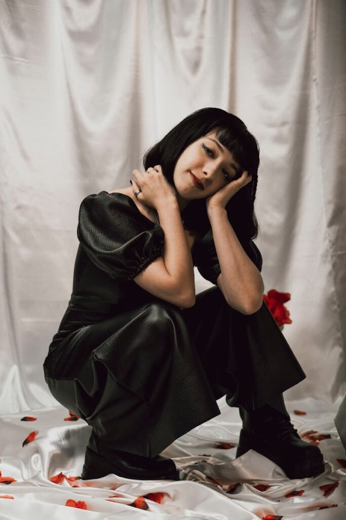 A stylish woman in a black gothic outfit poses gracefully with rose petals on a white silk backdrop.