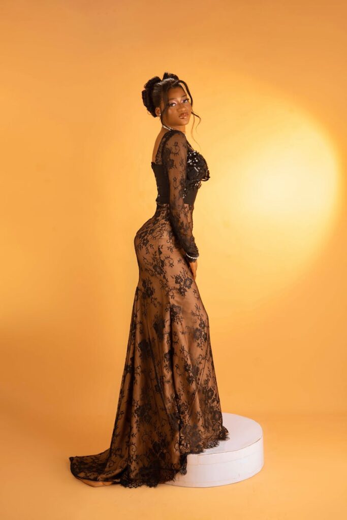 Stylish woman posing in a black lace gown against a vibrant yellow background in a studio setting.