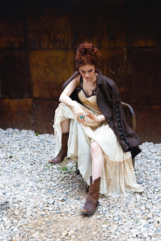 A woman in bohemian attire sits contemplatively outdoors on gravel.