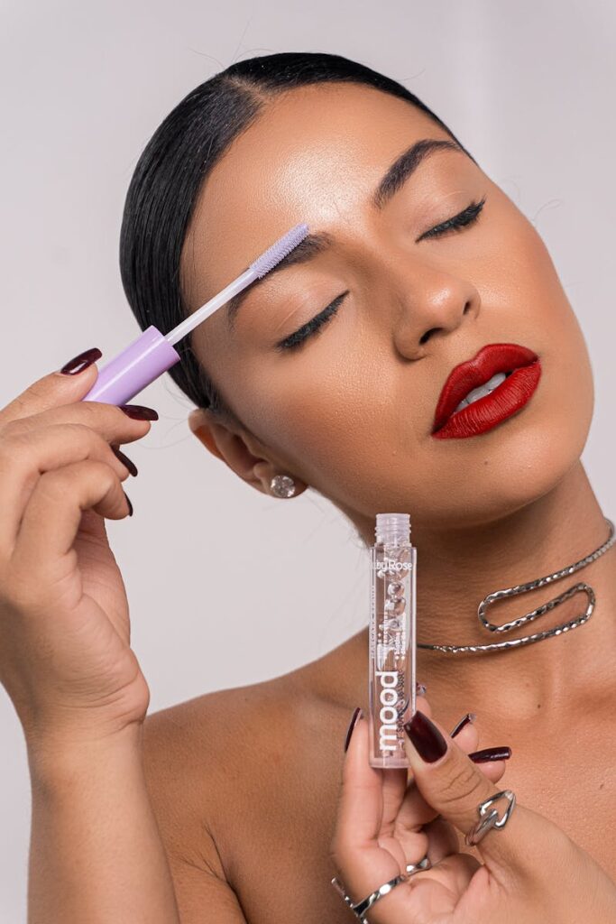 Woman with red lipstick applying eyebrow brush, showcasing flawless complexion.