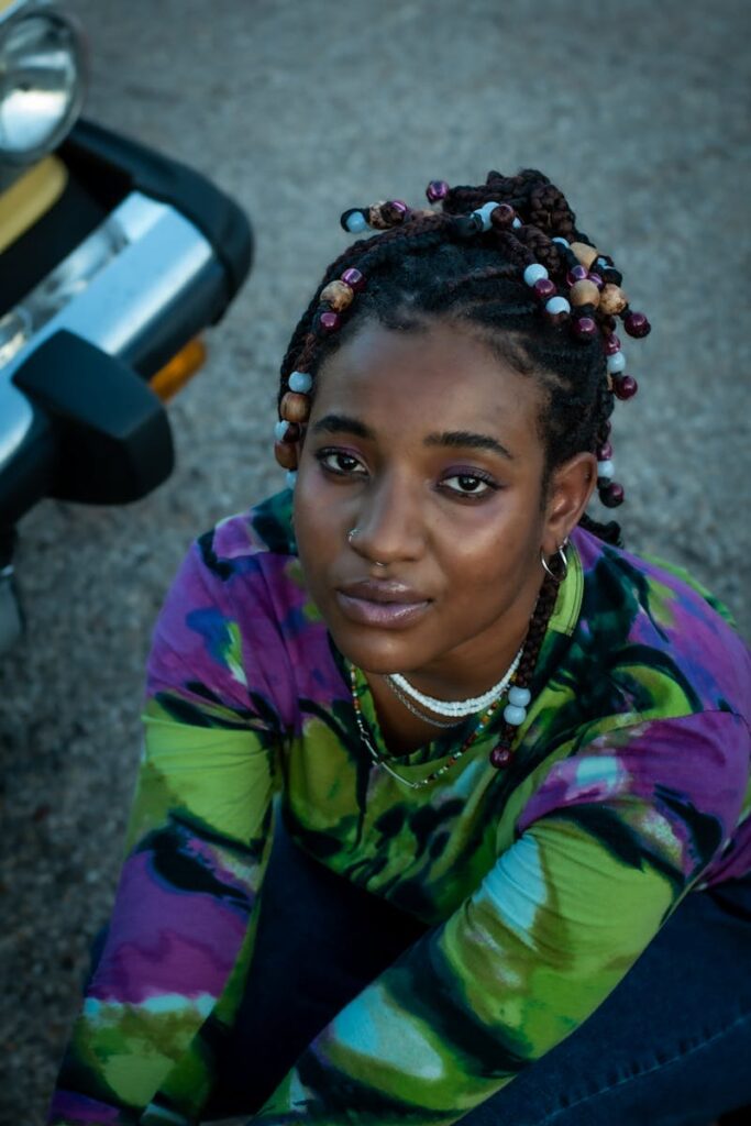 Young woman with bead-adorned braids sits near vehicle outdoors, striking pose.