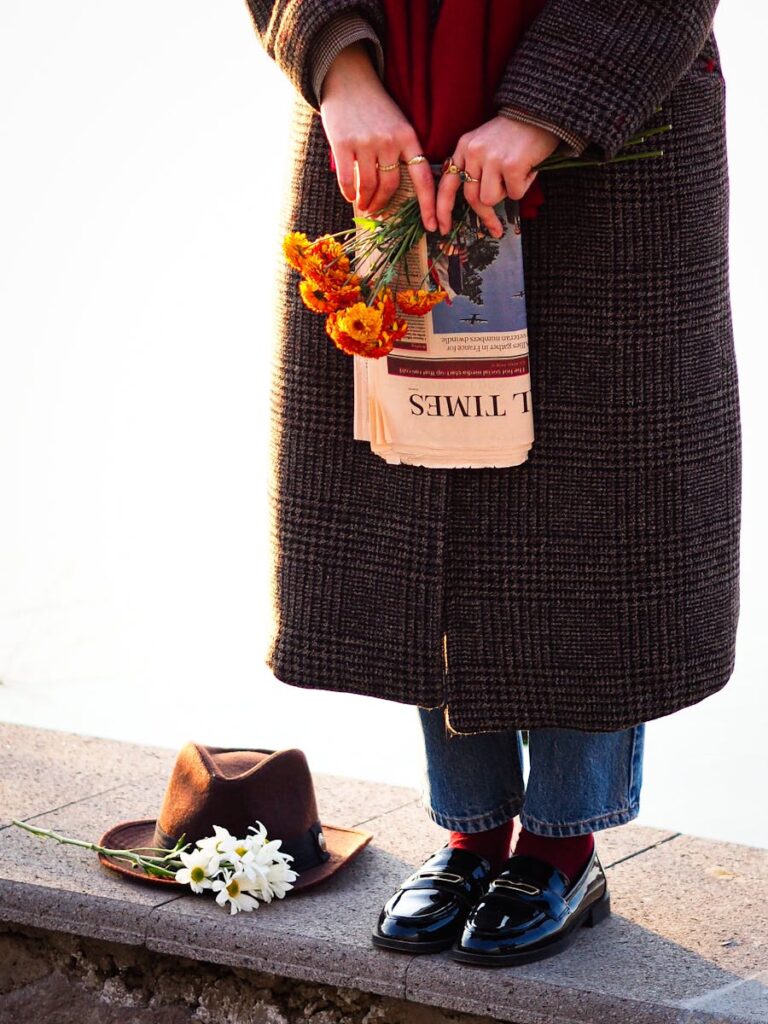 Woman in coat holds flowers and newspaper by the river, hat on stone ledge.