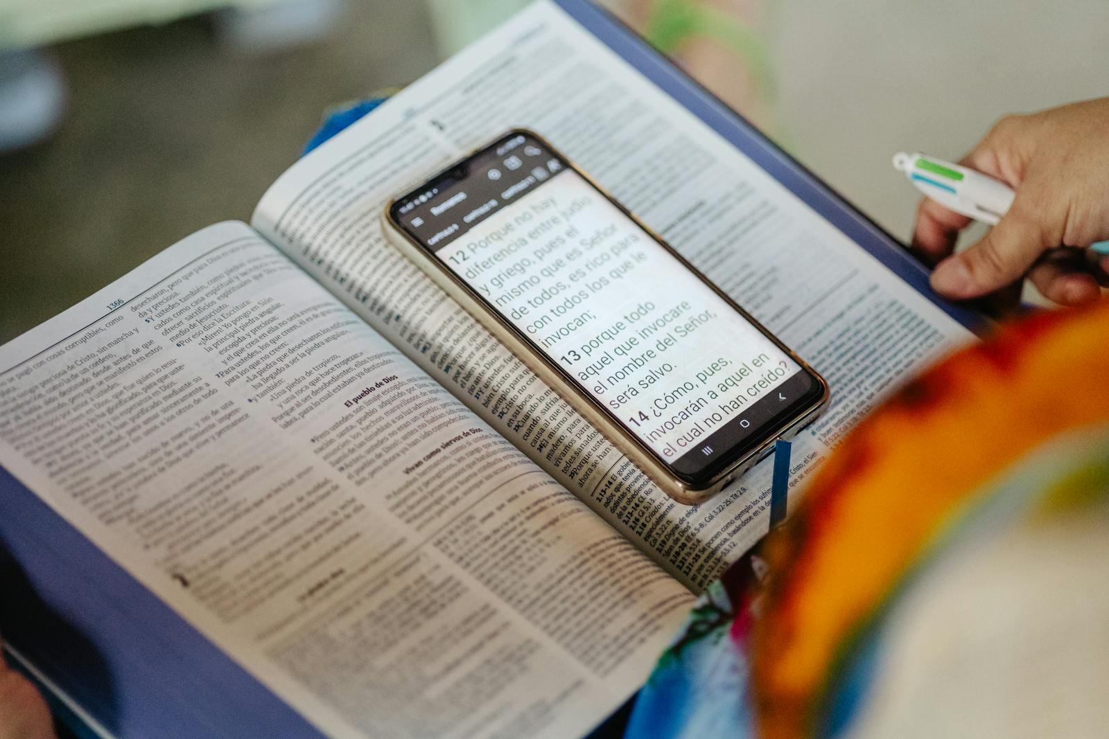 Open Bible with smartphone displaying scripture and hand holding marker.