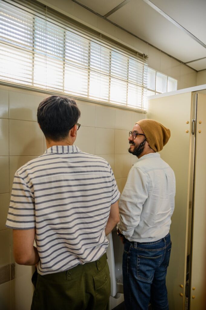 Two men wearing casual attire having a conversation in a restroom at urinals.