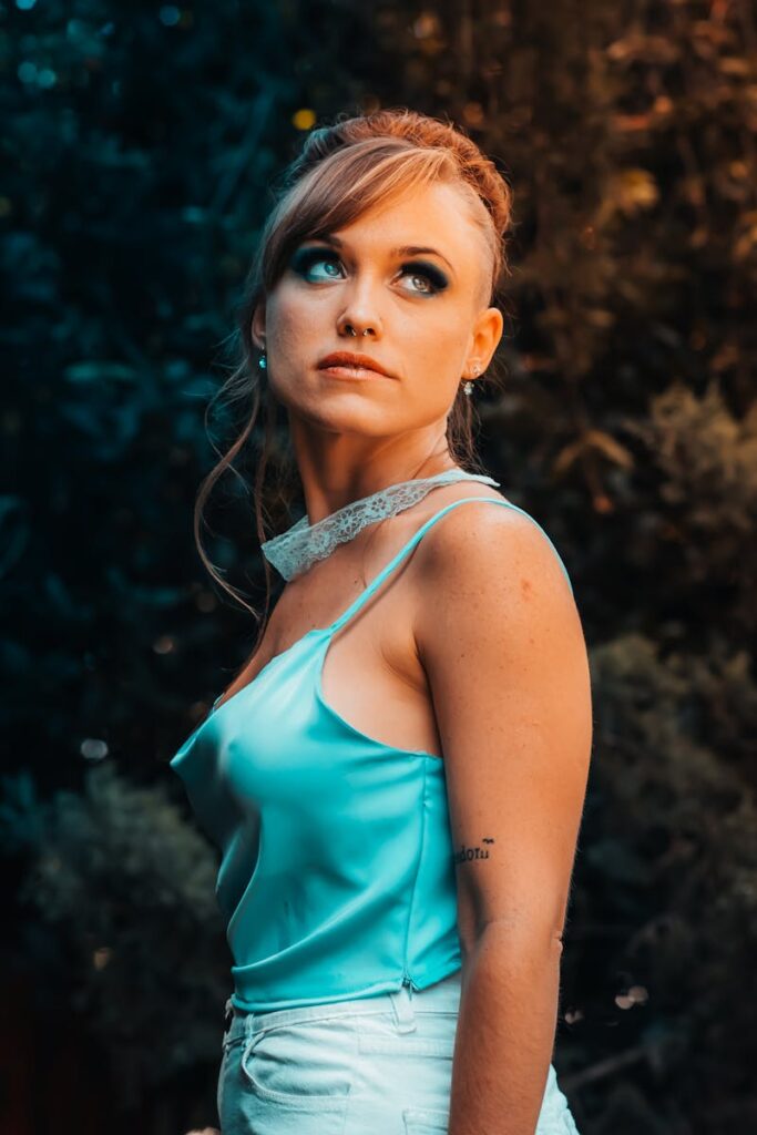 A contemplative woman in a blue satin top looks upwards in a lush outdoor setting.