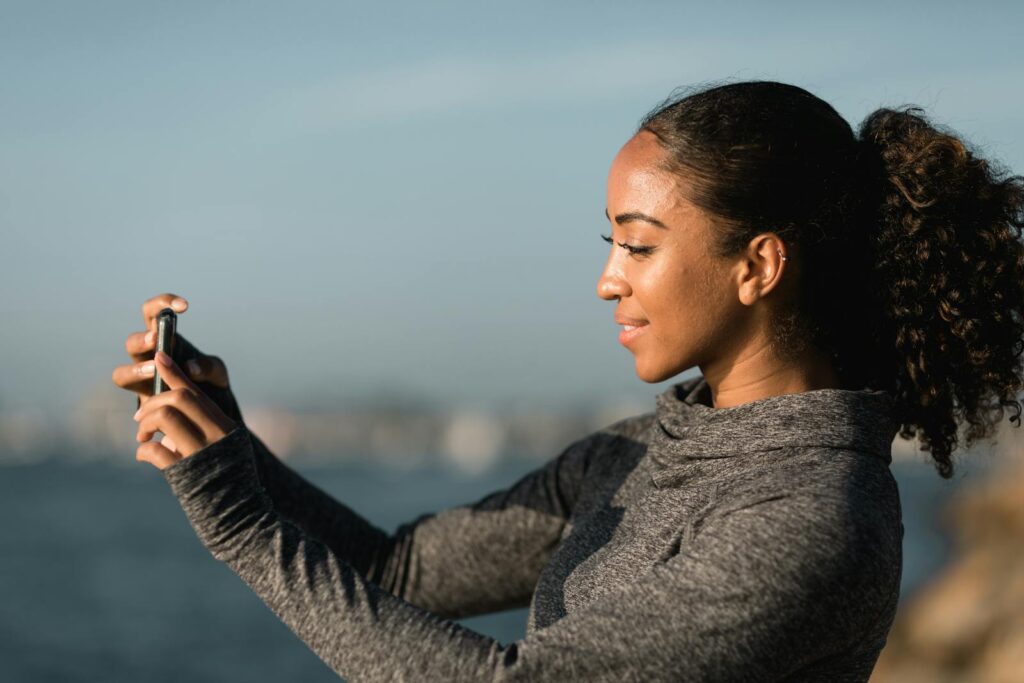 A young African American woman taking a selfie outdoors by the water on a sunny day.