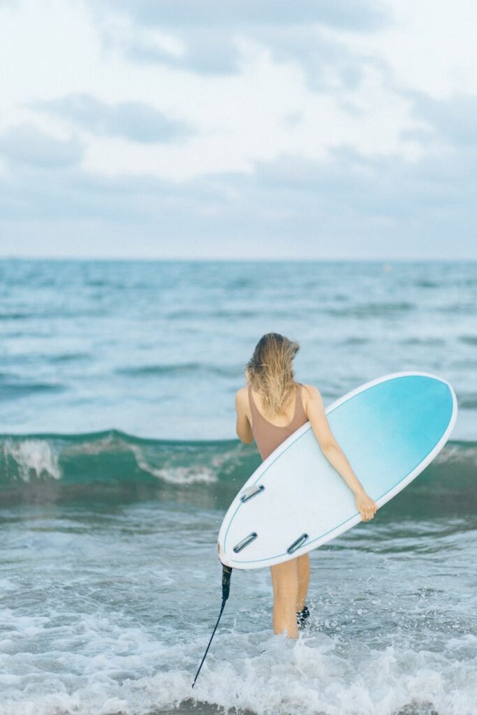 Woman carrying a surfboard, walking into the sea for surfing.