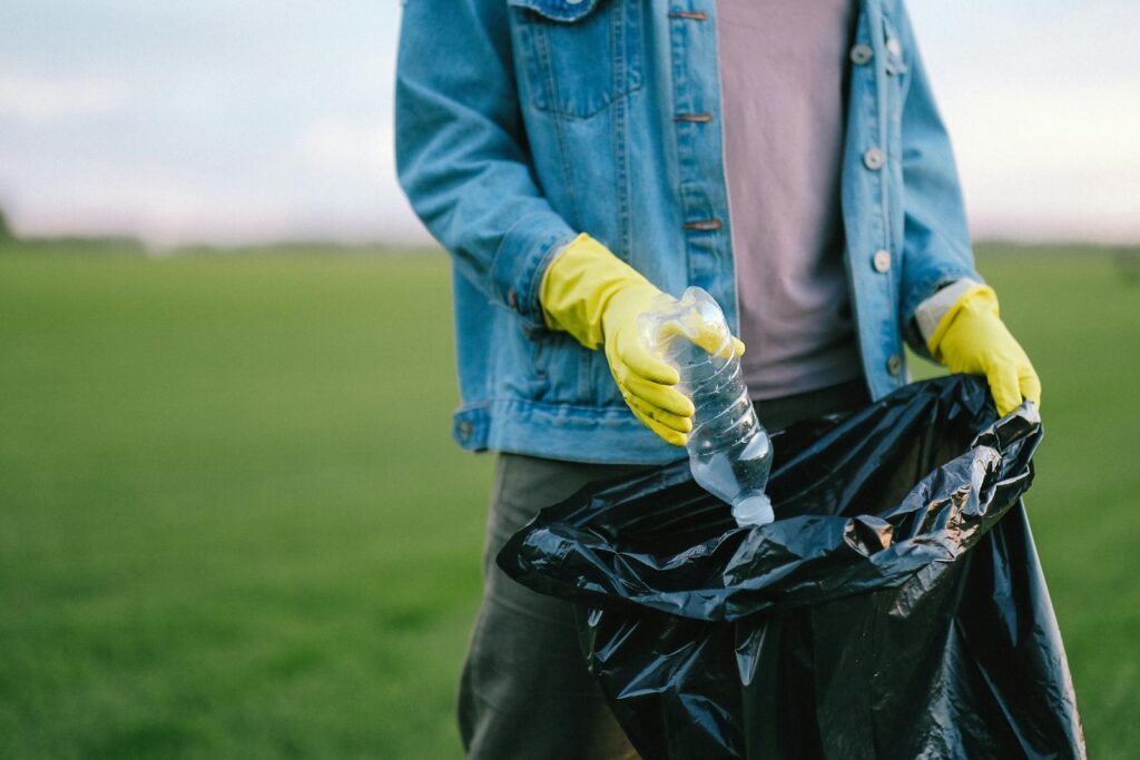 Volunteer in denim jacket and gloves picks up plastic bottle into trash bag in green field.