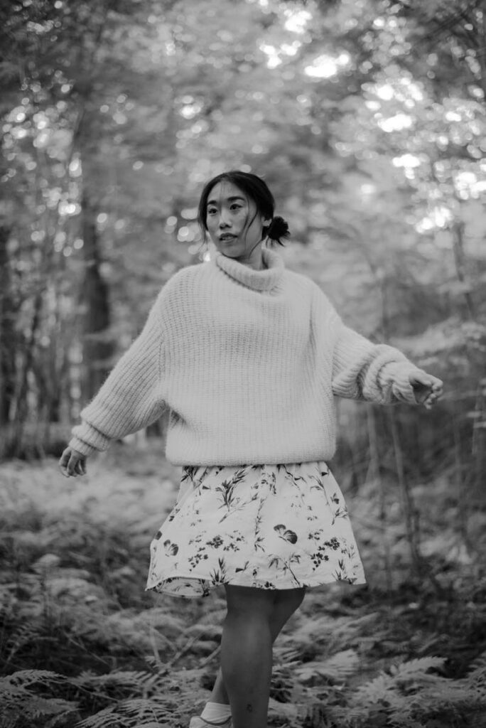 Black and white smiling Asian female in warm sweater and skirt resting in woods with lush plants