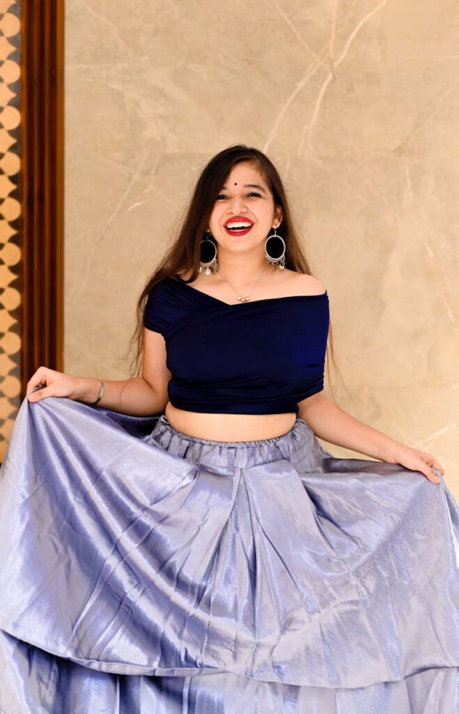 Smiling young Indian woman with long brown hair in stylish clothes holding long skirt in hands and standing near wall