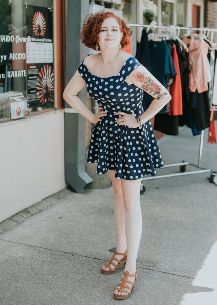 Stylish woman with tattoos in a polka dot dress posing confidently outdoors, exuding charm.