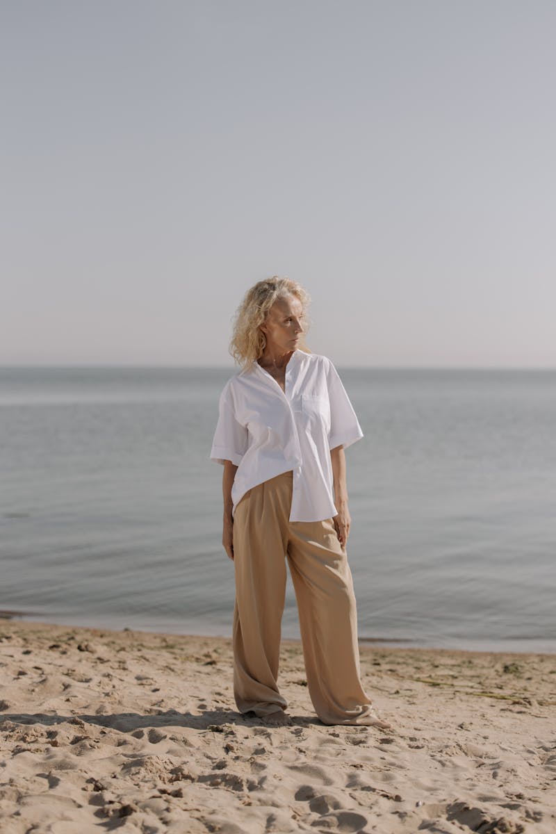 Elegant senior woman in white shirt standing on a tranquil beach by the sea.