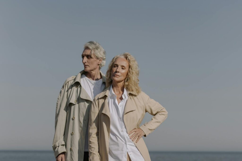 Stylish senior couple posing at the beach, under clear blue skies, wearing trench coats.