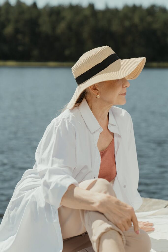 Senior woman in fashionable attire relaxing by a serene lake on a sunny day.