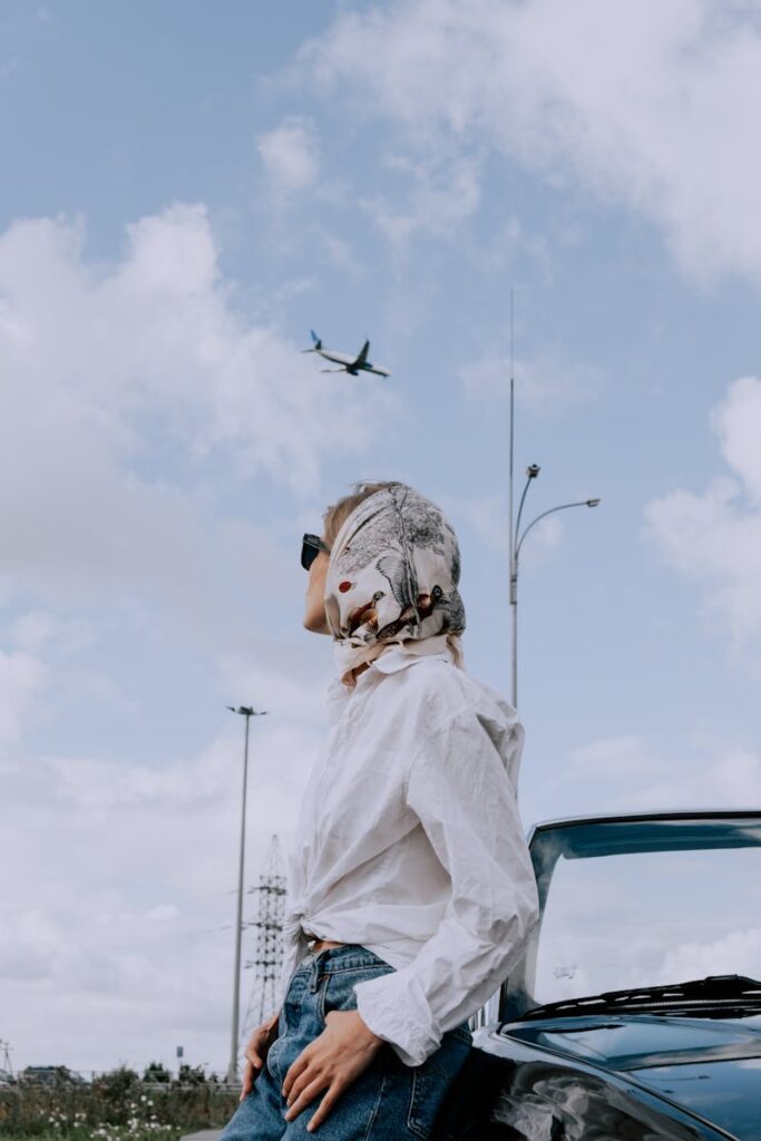 Woman in white top and denim leaning on car, wearing sunglasses and scarf, with airplane in cloudy sky.