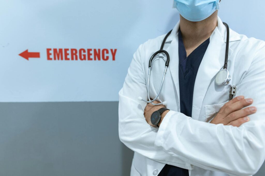 Healthcare worker in emergency room with stethoscope and facemask, cross-armed posture.