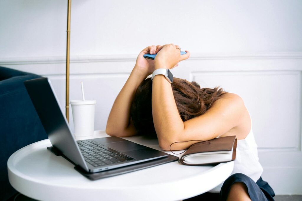 Young woman showing stress with laptop and phone at desk, embodying digital exhaustion.