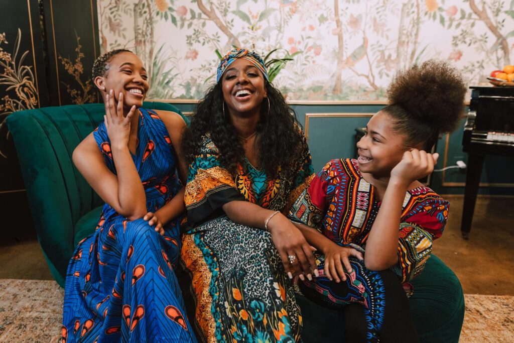 Three African American females joyfully celebrate Kwanzaa indoors with traditional attire.