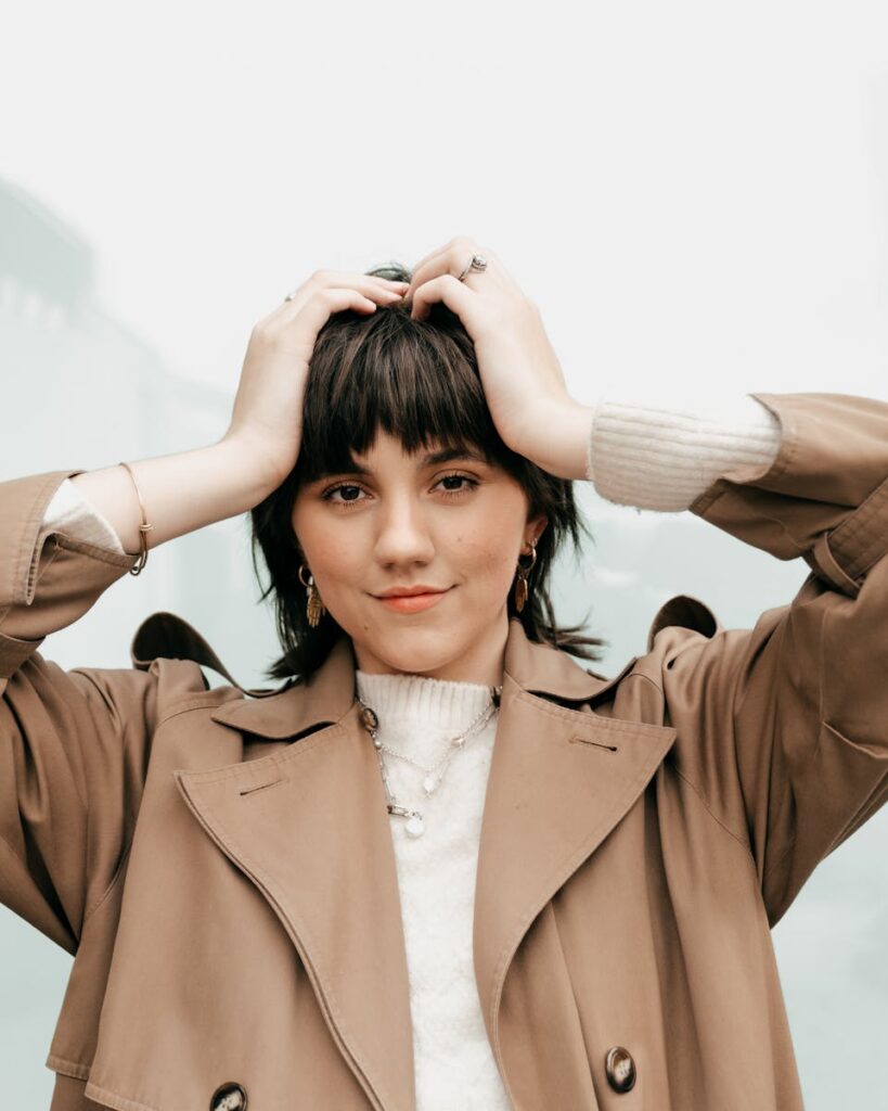 Calm young woman with short dark hair in stylish trench coat and outfits touching head and looking at camera against blurred light background