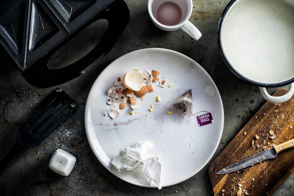 Chaotic breakfast setup with eggshells, tea tag, and cooking utensils on a kitchen counter.