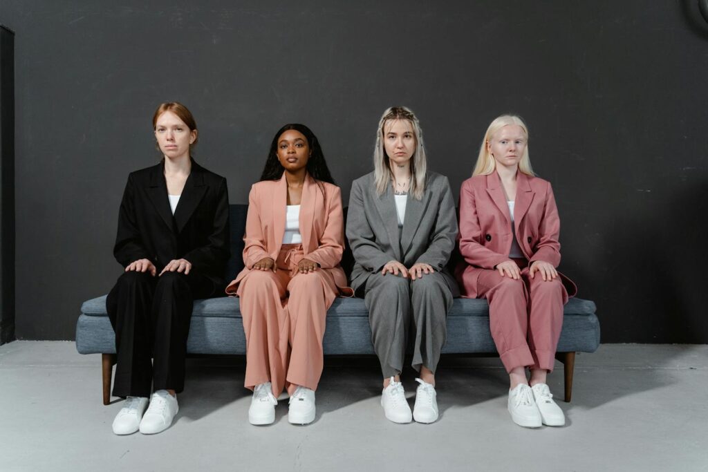 Four diverse women in stylish suits sitting on a couch in a studio setting.