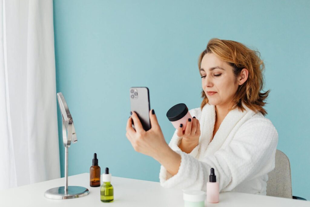 Woman in a white robe taking a selfie with skincare products, set against a light blue background.