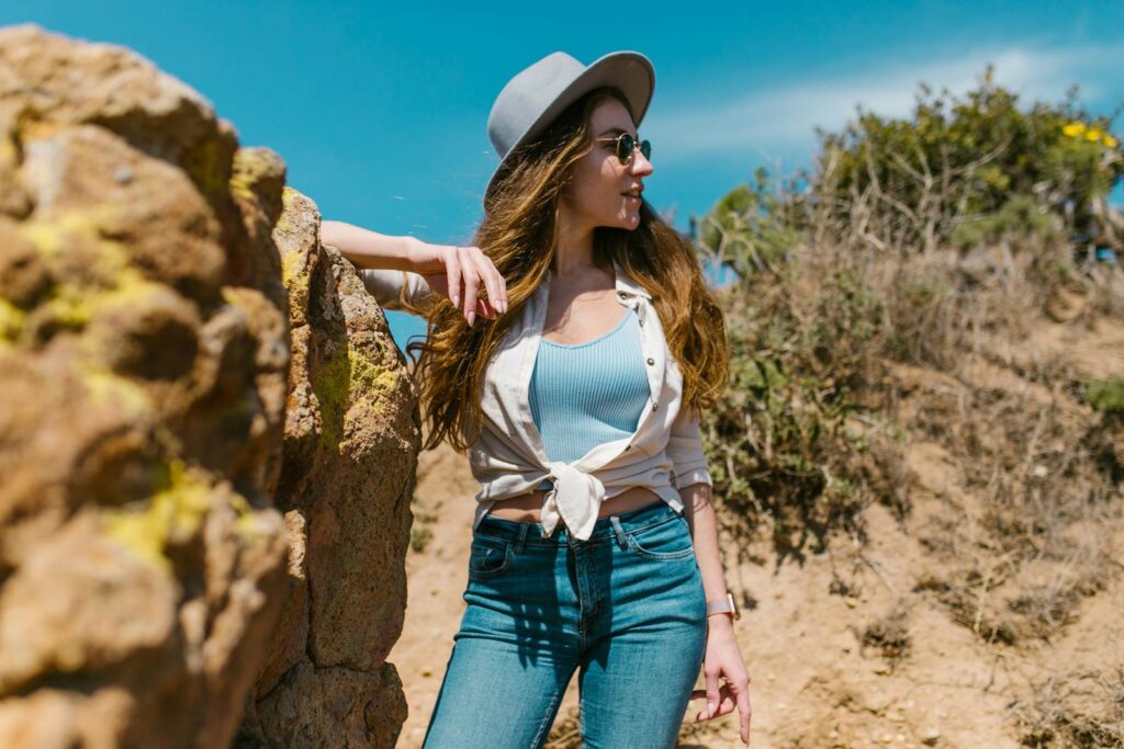 Stylish woman in jeans and hat posing by rocks outdoors on a sunny day.
