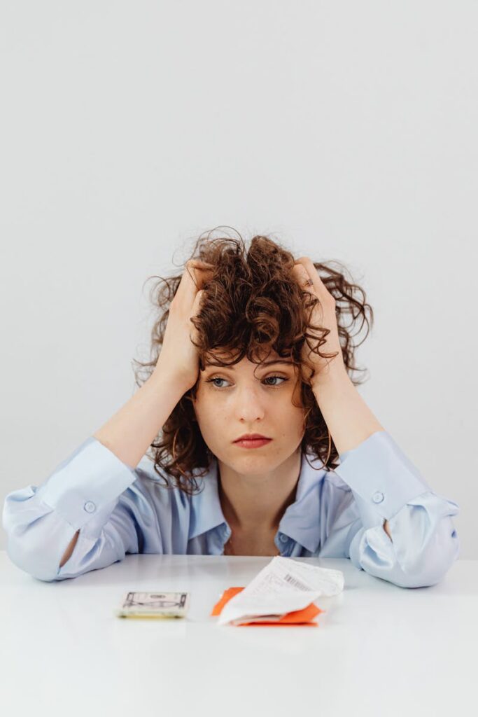 A worried woman with curly hair sits with her hands on her head surrounded by bills.