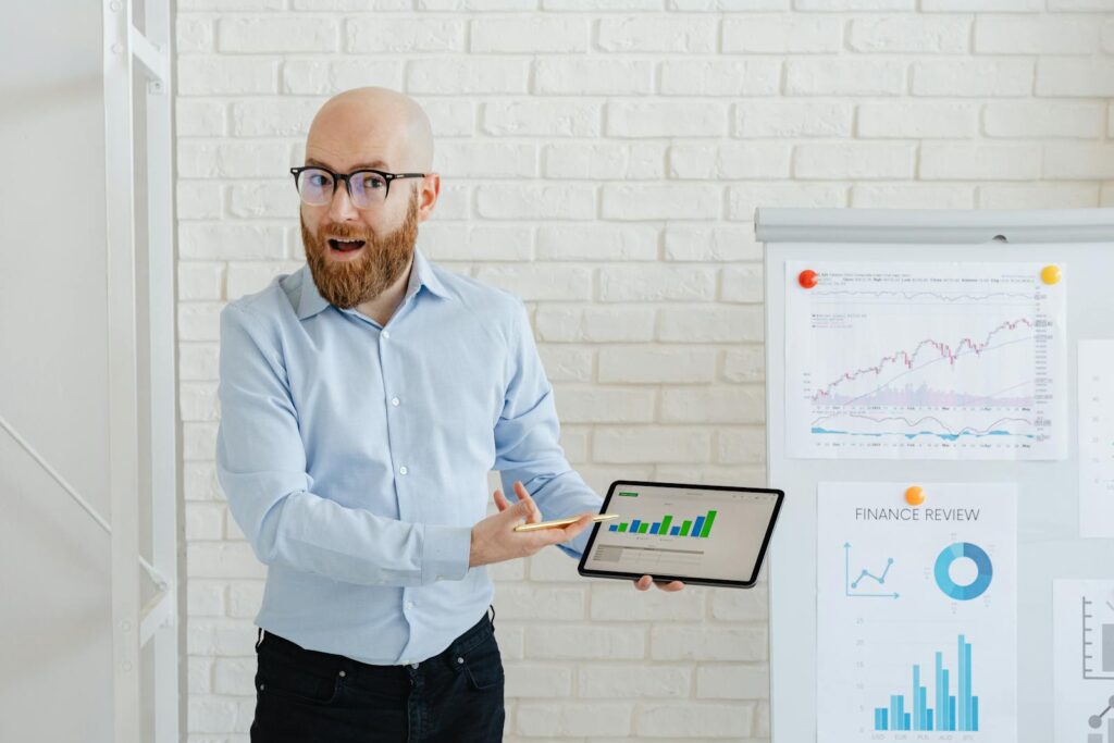 Bearded man in glasses presenting financial charts on tablet indoors.