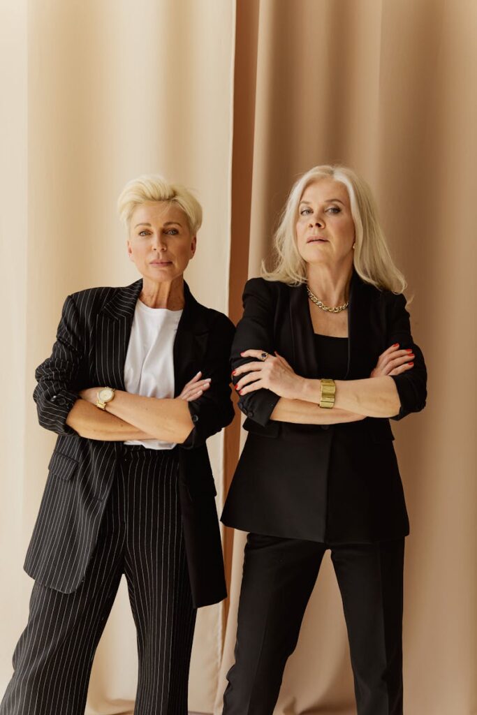 Two senior businesswomen pose confidently in stylish professional attire indoors.