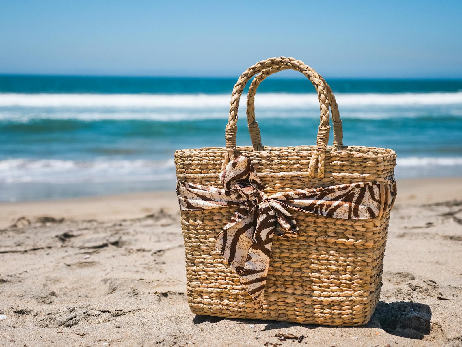 A stylish woven beach bag sits on a sunny beach with waves in the background.