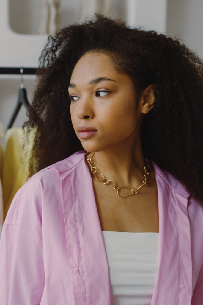 Elegant woman in a pink blouse with natural hair glances away, exuding stylish confidence indoors.
