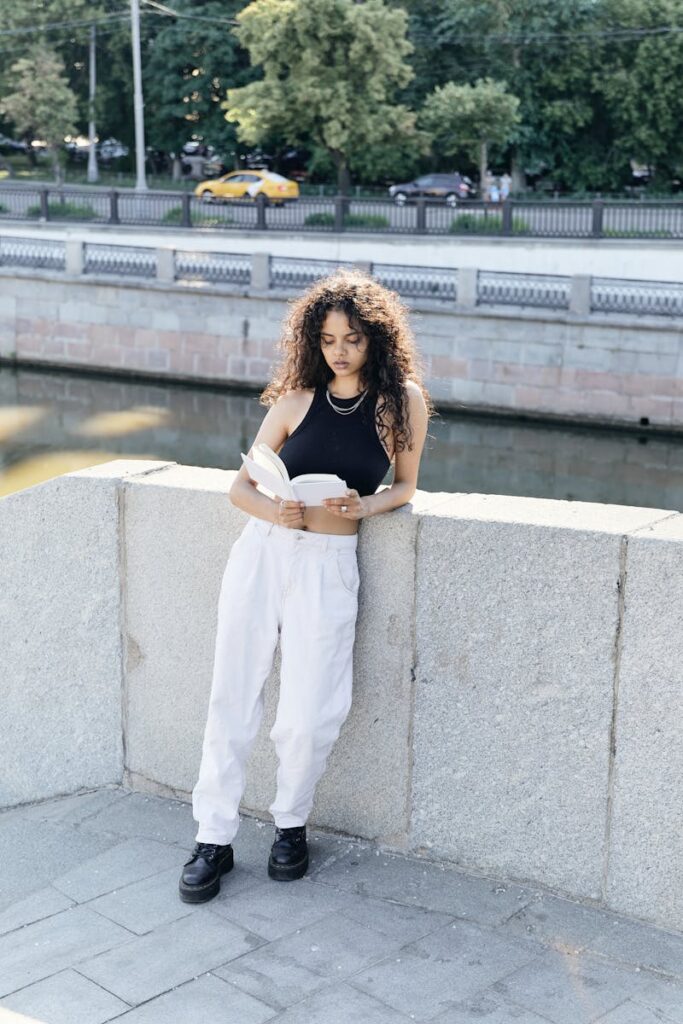 A fashionable young woman with curly hair reads by a stone railing on a sunny day.