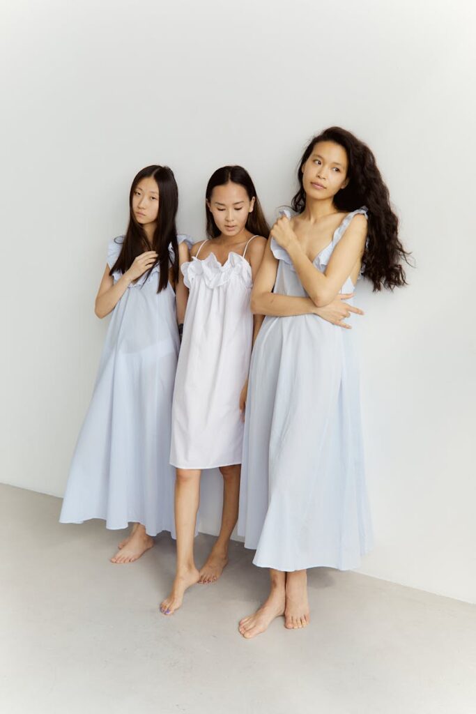 Three Asian women in light dresses posed gracefully indoors against a plain backdrop.