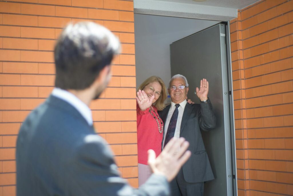 Smiling couple waving goodbye from home entrance with real estate agent in foreground.