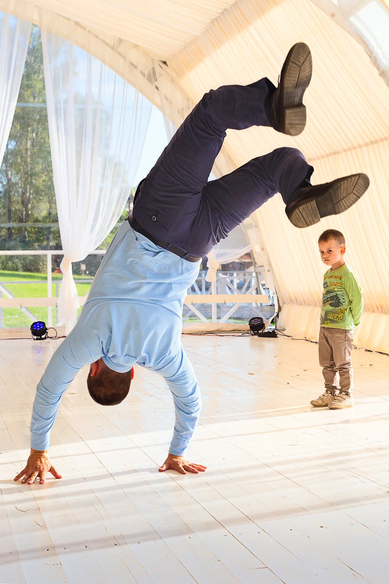 A father impresses his son by performing a handstand in a sunny indoor setting.