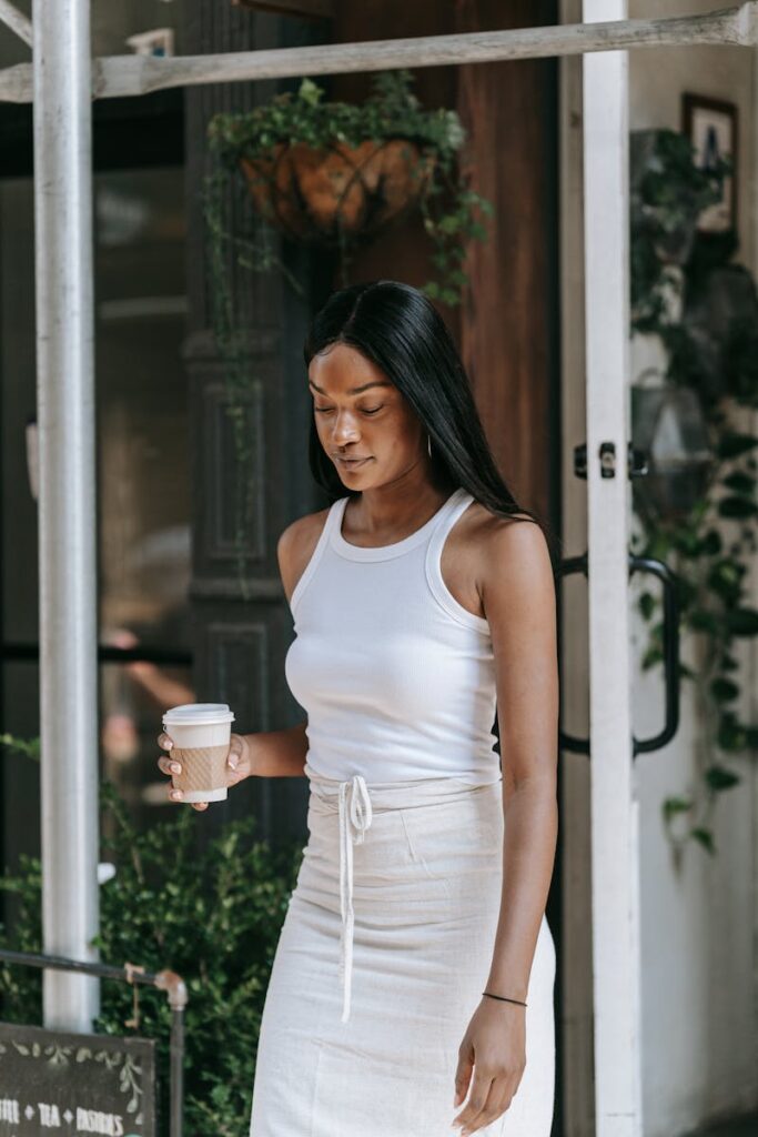 Stylish woman in white tank top and skirt holding coffee while walking outside.