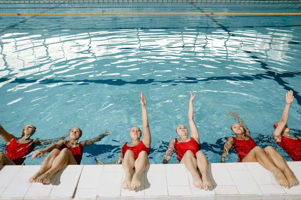 Team of women in red swim caps and costumes practicing synchronized swimming by the pool.