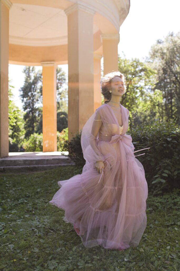 A woman in a pink sheer dress gracefully walks near a gazebo in a sunlit garden.