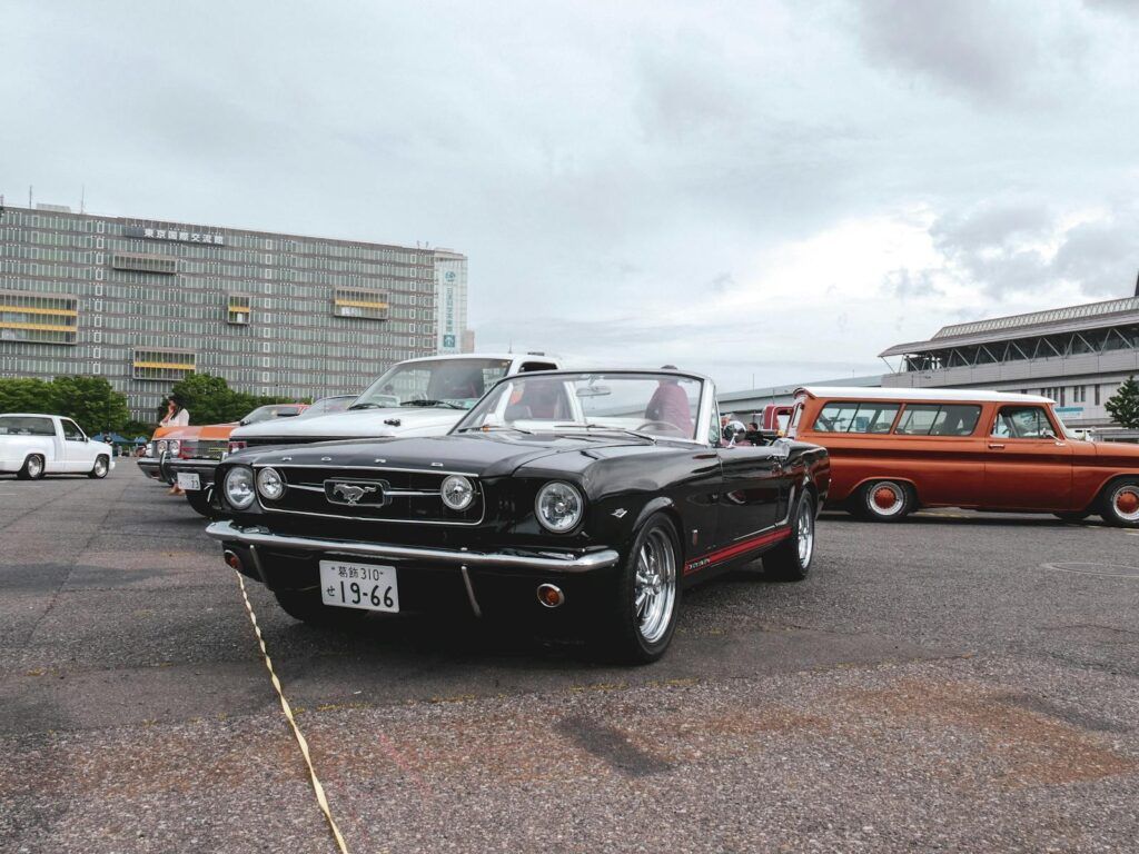 A classic black Ford Mustang at a vintage car meetup with an urban backdrop.